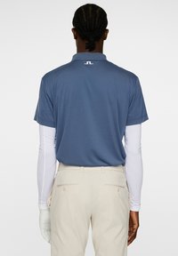 Miestenská golfová polokošeľa s krátkym rukávom v modrej farbe s golierom a logom na zadnej strane. Svetlo béžové nohavice s vreckami a jemnou textúrou. Biela košeľa s dlhým rukávom pod tým.