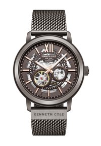 Montre automatique avec un bracelet en maille, accents bronze et un cadran transparent dévoilant des mécanismes intérieurs complexes. Comprend des index horaires et le logo de la marque.