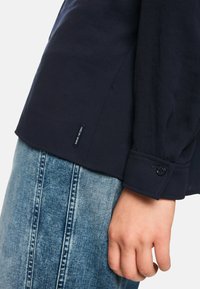 Navy long-sleeve shirt with buttoned cuff, paired with blue denim. Tag visible at hem. Smooth texture, classic fit, casual design.