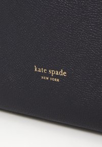 Juodas tekstūruotas oda su matomu auksiniu "kate spade NEW YORK" logotipu. Paviršius yra šiek tiek akmenuotas, kas rodo patvarumą ir kokybę.