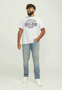 Smiling man with short dreadlocks wearing white Jack & Jones t-shirt, light blue jeans, and white sneakers, standing with one hand in pocket.