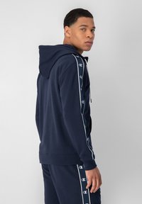 Felpa con cappuccio blu navy con strisce laterali bianche e loghi. Tessuto morbido, maniche lunghe, tasca a marsupio frontale e cappuccio con cordino.