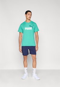 Τιρκουάζ μπλουζάκι PUMA με μεγάλο λευκό λογότυπο, συνδυασμένο με ναυτικό σορτς. Το μοντέλο φορά λευκά αθλητικά παπούτσια και κάλτσες PUMA.