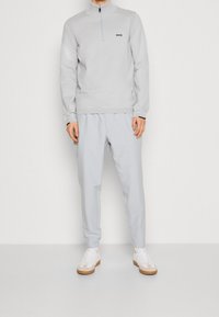 Sweatshirt zippé gris clair avec un logo, associé à un pantalon tapered gris clair assorti et des baskets blanches avec des semelles marron.