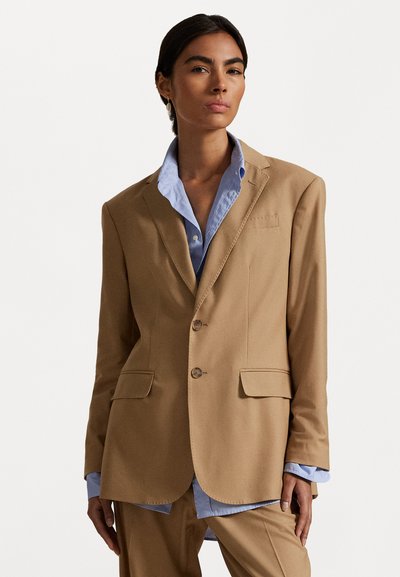 Blazer beige en tissu lisse avec deux poches avant, trois boutons et un col cranté, porté sur une chemise bleu clair.