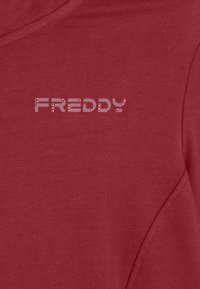 T-shirt en coton rouge avec un texte graphique "FREDDY" dans un design contrasté. Le tissu semble lisse avec un léger éclat.