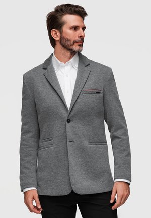 Mand med skæg iført grå blazer over hvid skjorte, kigger mod venstre foran en ensfarvet lys baggrund.