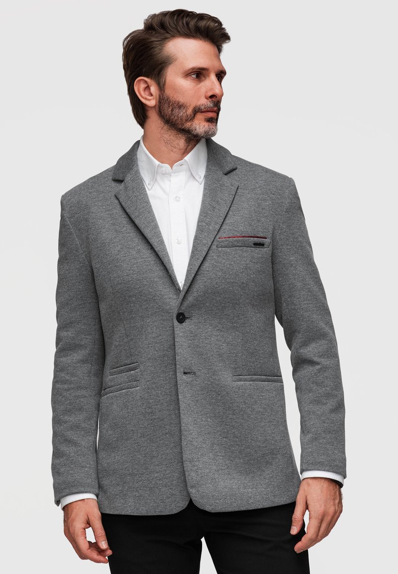 Homme à la barbe portant un blazer gris sur une chemise blanche, regardant vers la gauche sur un fond clair uni.