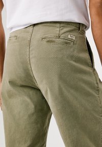 Close-up de uma pessoa a usar calças verde-azeitona com bolsos traseiros e uma camisa branca metida para dentro.