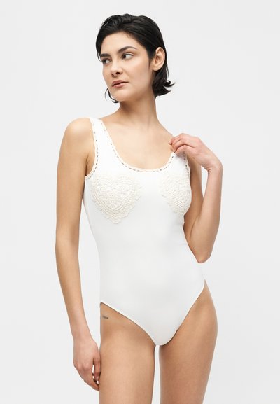 Γυναίκα με κοντά σκουρόχρωμα μαλλιά, φοράει ένα λευκό αμάνικο κορμάκι με σχέδια καρδιάς από πλέξη στο στήθος, κοιτάζοντας πλαγίως.