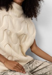 Gruby sweter w kolorze kremowym z wysokim dekoltem, z fakturowanym wzorem fal. Połączony z jasnymi spodniami w marmurkowy wzór.