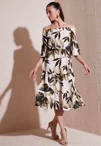 Off-the-shoulder dress featuring a floral print in green, blue, and beige, with a flared hem and textured fabric. Model wears beige heels.