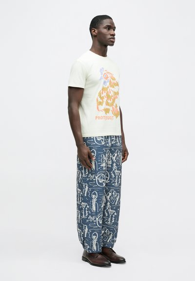 Φωτεινό πράσινο μπλουζάκι με πορτοκαλί γραφικά και μπλε λεπτομέρειες, συνδυασμένο με μπλε παντελόνια φαρδιάς γραμμής με σχέδια και λευκές εικονογραφήσεις, καθώς και σκούρα παπούτσια.