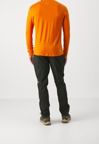 Pitkähiha tshirt oranssina, mustat housut taskuilla ja ruskeat vaelluskengät. T-paidassa on yksinkertainen muotoilu ja housuissa on teksturoitu pinta.