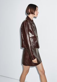 Frau mit kurzen braunen Haaren, die vor einer weißen Wand entlanggeht, trägt eine dunkelbraune glänzende Lederjacke und einen passenden kurzen Rock.