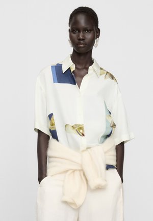 Mannequin portant une chemise blanche à manches courtes avec des imprimés bleus et jaunes et un pull couleur crème noué autour de la taille.