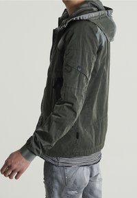 Lekka kurtka w oliwkowym odcieniu, z zamkiem z przodu, kapturem i teksturowanymi akcentami. Posiada szare wykończenie oraz guzikowe zdobienie na rękawach.