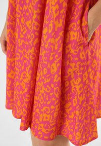 Lyhyt mekko, jossa on eloisa vaaleanpunainen ja oranssi leopardikuvio, pehmeä rakenne ja sivutaskut. Väljä helma ja rento muotoilu.