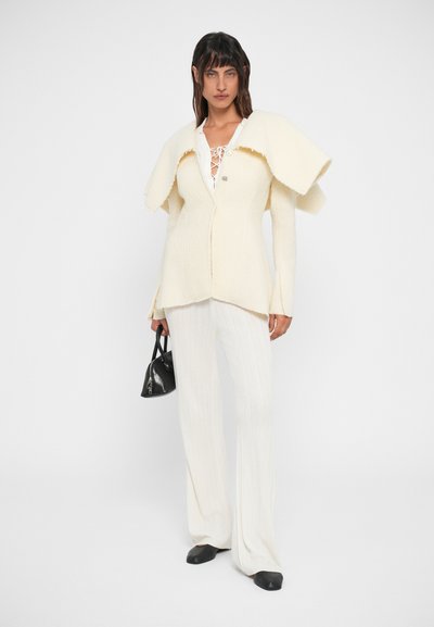 Blazer creme com textura e gola oversized, combinado com calças brancas caneladas e sapatos pretos rasos, segurando uma pequena bolsa preta.