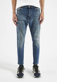 Jeans de denim azul con un ajuste slim, detalles desgastados y textura desvanecida. Cuenta con cinco bolsillos y cierre de botón. Llevado con zapatillas deportivas.