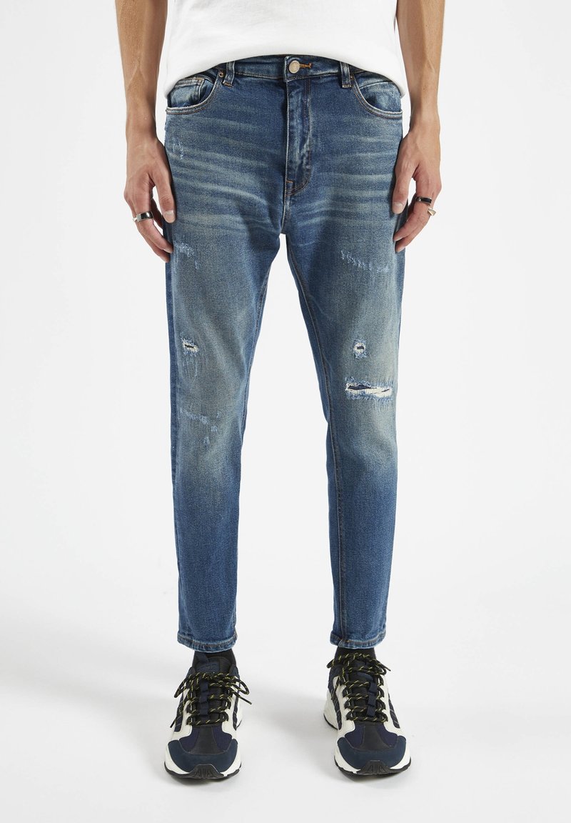 Jeans de denim azul con un ajuste slim, detalles desgastados y textura desvanecida. Cuenta con cinco bolsillos y cierre de botón. Llevado con zapatillas deportivas.