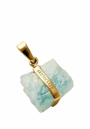Colgante de oro que sostiene una piedra preciosa rugosa de color azul claro con "ARISTOCRAZY" grabado en la correa de metal.