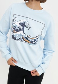 Sudadera azul cielo con un estampado gráfico de olas del océano. Presenta un cuello y puños acanalados, y un corte relajado diseñado para la comodidad.