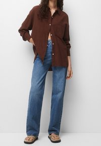 Chemise brune à boutons avec des manches longues et une poche poitrine, assortie à un jean droit bleu clair et des tongs beiges.