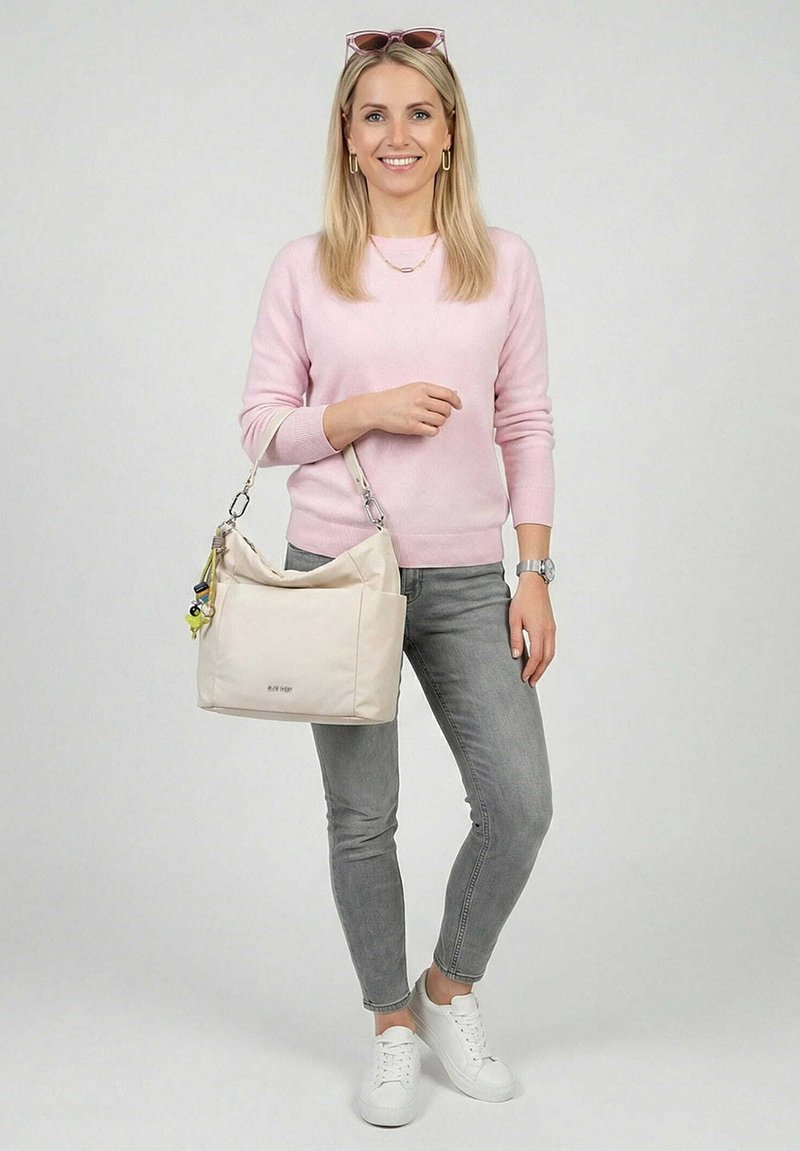 Mujer con suéter rosa y jeans grises sostiene un bolso beige con llavero, sonriendo, con gafas de sol en la cabeza contra un fondo liso.
