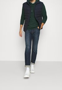 Granatowa pikowana kamizelka na zielonej bluzie z kapturem, do tego podwinięte ciemne jeansy i białe trampki. Strój ma luźny krój i casualowy styl.