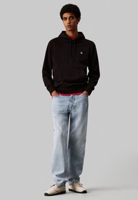 Felpa nera con tasca frontale e cappuccio, indossata sopra una maglietta rossa. Jeans azzurri a vestibilità rilassata e sneakers beige completano il look.