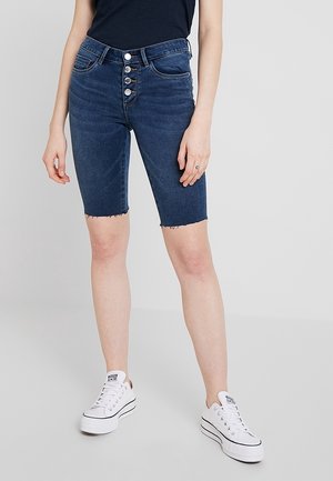 Frau trägt dunkelblaue, hoch taillierte Jeansshorts mit Knopfleiste und weiße Sneaker, steht vor einem schlichten hellen Hintergrund.