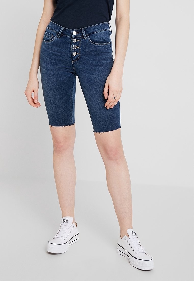 Femme portant un short en jean bleu marine taille haute avec boutons sur le devant et des baskets blanches, debout devant un fond clair uni.