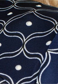 Laikainen kangas, jossa on valkoisia aaltoilevia viivoja ja pilkullisia yksityiskohtia. Pintakuvio on texturoitu, rakenteellinen muotoilu ja kontrastivärinen brodeeraus.