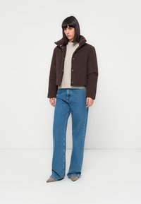Donna in piedi contro un muro bianco, vestita con una giacca imbottita marrone scuro, un maglione beige, jeans larghi blu e scarpe grigie a punta.