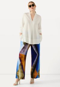 Witte blouse met lange mouwen en een V-halslijn, voorzien van twee zijzakken, gecombineerd met kleurrijke, geometrisch patroon wijdlopende broek. Beige sandalen met bandjes.
