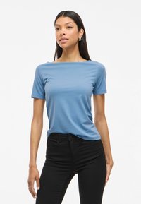 Blå kortärmad topp med vid rundad halsringning och slät, lätt tyg, matchad med svarta åtsittande jeans. Designen är enkel och avslappnad.