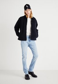 Svart denimjacka över en vit t-shirt, ljusblå jeans och svarta skor. Kappen är svart med vita logotypdetaljer.