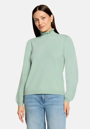 Woman wearing light green ruffled mock neck sweater and light blue jeans, standing against a plain light background.
