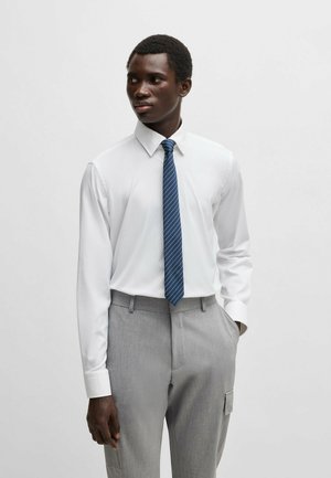 Chemise blanche à manches longues associée à un pantalon gris avec une poche latérale. La chemise a un col standard et la cravate est bleue avec des rayures.
