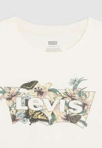 Tricou de culoare crem cu un design floral și fluturi, având logo-ul "Levi's" în litere albe îngroșate la centru.