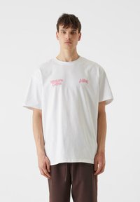 Biela bavlnená tričko s krátkymi rukávmi, s ružovými potlačami na hrudi. Strih je voľný s okrúhlym výstrihom.