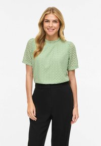 Haut vert clair à manches courtes avec motif festonné texturé et encolure à volants, associé à un pantalon noir ajusté.