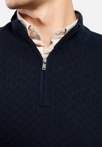 Maglione zip-up blu scuro in tessuto testurizzato, con un colletto alto e una piccola zip. Sotto, è visibile una camicia con colletto di colore crema.