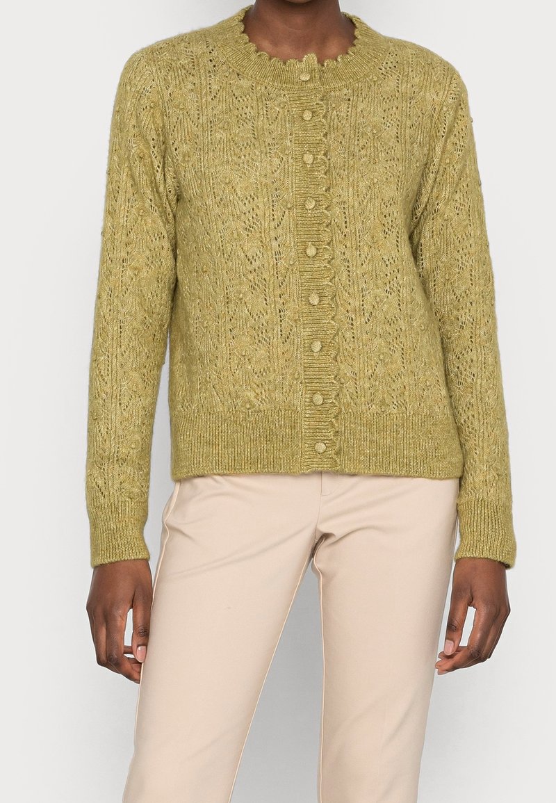 Woman wearing a knitted olive green buttoned cardigan with intricate patterns and beige pants, standing against a plain background.