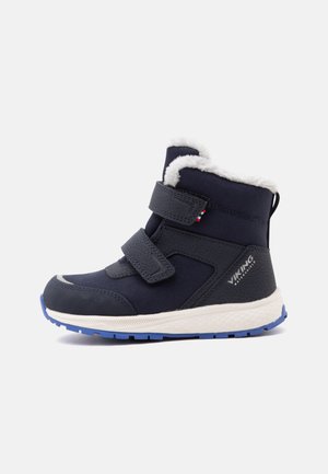 Bota de invierno para niños azul marino con forro de piel blanca, dos tiras de velcro y suela de goma azul, etiquetada como impermeable en el talón.