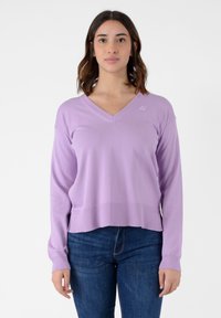 Maglione viola chiaro con scollo a V, maniche lunghe, orlo a coste e dettaglio logo sul petto. Realizzato in un tessuto morbido e elasticizzato. Indossato con jeans.