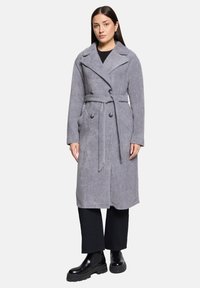 Manteau gris ceinturé à double boutonnage, larges revers, tissu texturé et poches latérales, assorti à un pantalon noir et des bottes.