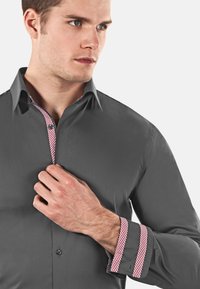 Camicia grigia con un colletto e polsini interni a motivo a quadri rosso e bianco in contrasto. Il tessuto sembra liscio, con un design aderente.