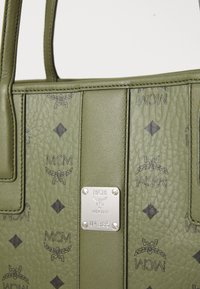 Sac en cuir vert olive MCM avec un motif de logo noir et une plaque en métal argenté affichant le nom de la marque et le numéro de série U4385.